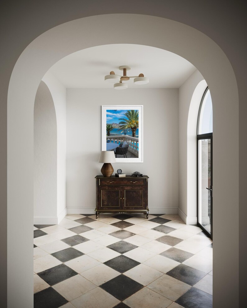 May include: An interior hallway with a black and white checkered floor, leading to a framed picture of a tropical scene. A dark wood cabinet with a lamp and a clock sits below the picture. The hallway is framed by white archways.