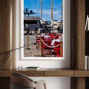 May include: A framed photograph of a harbor scene with yachts and sailboats under a bright blue sky. Red chairs and tables with white tablecloths are set up on a cobblestone street, suggesting a restaurant or cafe.