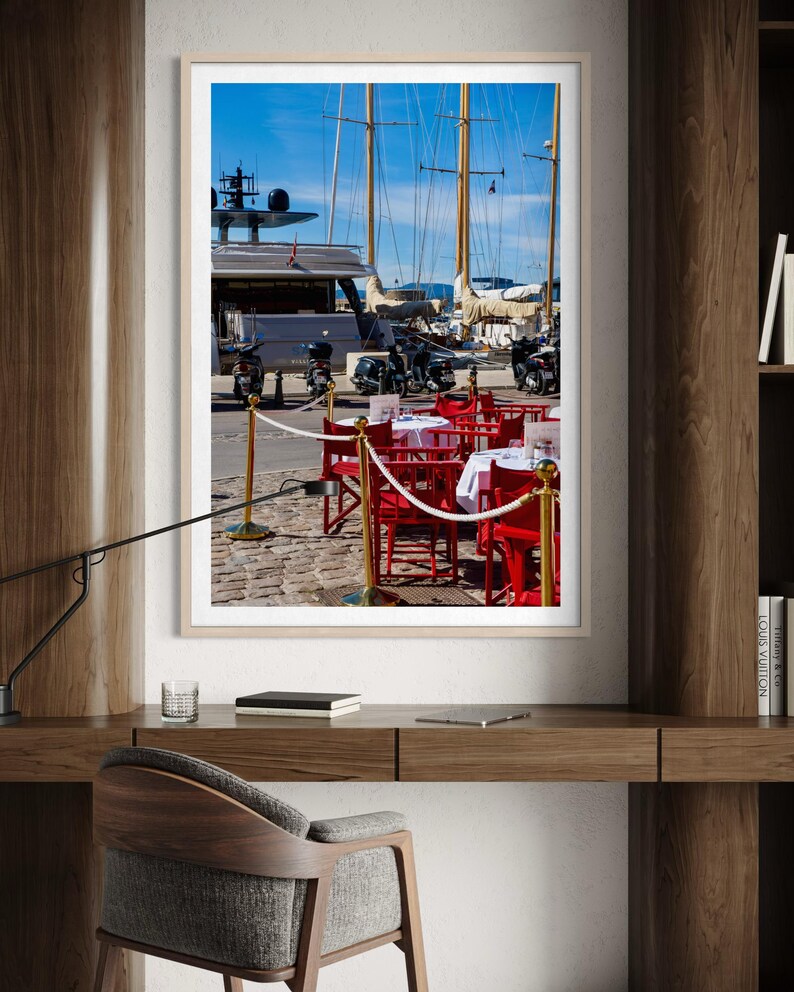 May include: A framed photograph of a harbor scene with yachts and sailboats under a bright blue sky. Red chairs and tables with white tablecloths are set up on a cobblestone street, suggesting a restaurant or cafe.