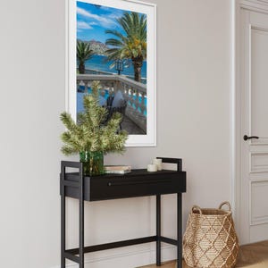 May include: A black console table with a drawer, topped with a green glass vase holding greenery, books, and candles. A framed picture of a tropical scene hangs above the table. A woven basket sits on the floor.