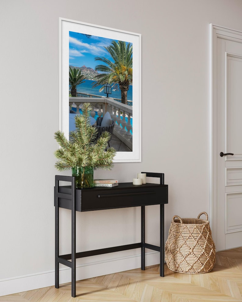 May include: A black console table with a drawer, topped with a green glass vase holding greenery, books, and candles. A framed picture of a tropical scene hangs above the table. A woven basket sits on the floor.
