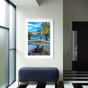 May include: A framed photograph of a tropical scene with palm trees, a blue sky, and a view of the ocean. The photograph is displayed on a white wall above a blue velvet bench with gold legs. The floor has a black and white striped pattern.