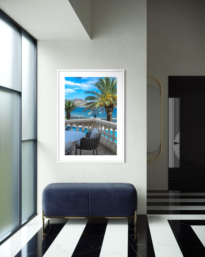 May include: A framed photograph of a tropical scene with palm trees, a blue sky, and a view of the ocean. The photograph is displayed on a white wall above a blue velvet bench with gold legs. The floor has a black and white striped pattern.