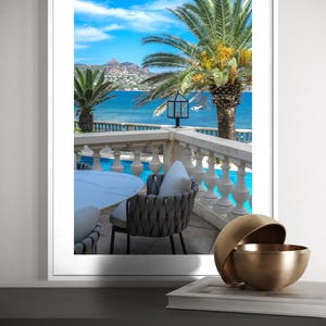 May include: A framed print of a coastal scene featuring a balcony with a table and chairs overlooking the ocean. Palm trees frame the view of the blue water and sky. A decorative gold bowl sits on a table in the foreground.