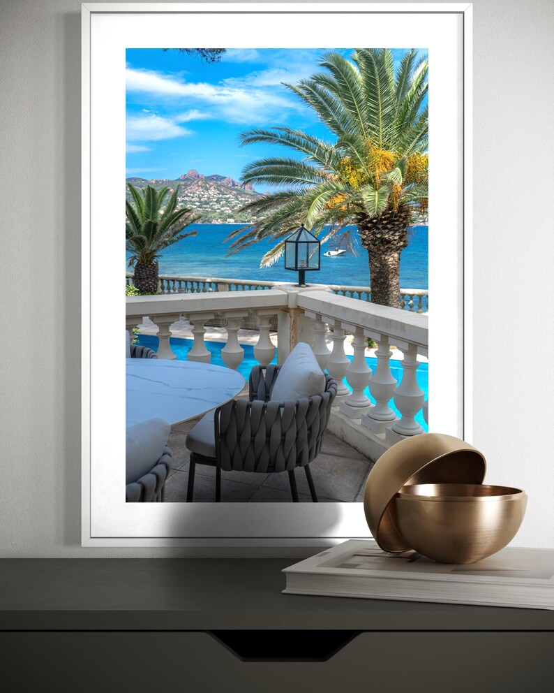 May include: A framed print of a coastal scene featuring a balcony with a table and chairs overlooking the ocean. Palm trees frame the view of the blue water and sky. A decorative gold bowl sits on a table in the foreground.