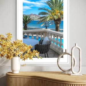 May include: A framed print of a scenic coastal view. The image features palm trees, a blue ocean, and a white balcony with a table and chair. A vase of yellow flowers and decorative sculptures sit on a wooden cabinet in the foreground.