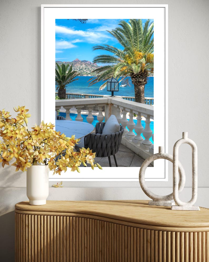 May include: A framed print of a scenic coastal view. The image features palm trees, a blue ocean, and a white balcony with a table and chair. A vase of yellow flowers and decorative sculptures sit on a wooden cabinet in the foreground.