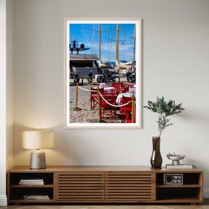 May include: A framed photograph of a harbor scene. Red chairs and tables with white tablecloths are set up on a cobblestone street. Sailboats and motorcycles are visible in the background under a bright blue sky.