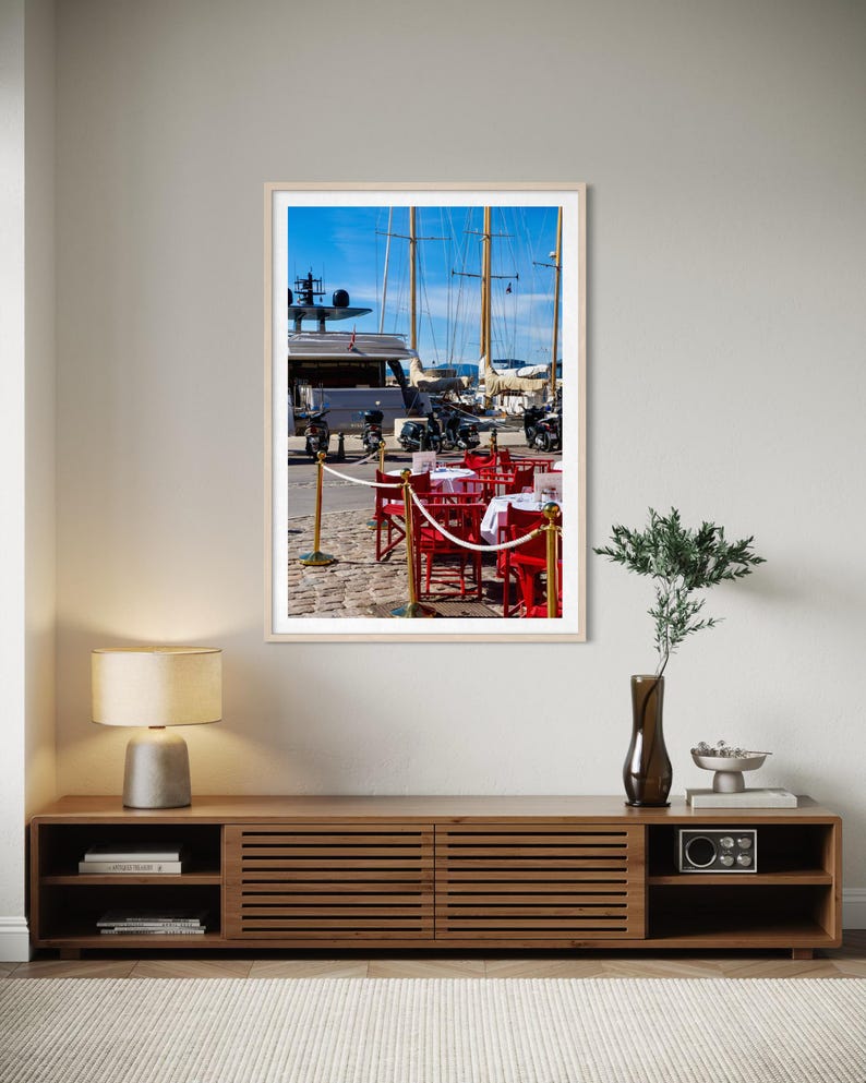 May include: A framed photograph of a harbor scene. Red chairs and tables with white tablecloths are set up on a cobblestone street. Sailboats and motorcycles are visible in the background under a bright blue sky.