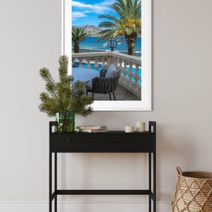 May include: A framed photograph of a scenic coastal view with palm trees, a pool, and a table and chair set. A black console table with a green vase and candles is in the foreground. A woven basket sits on the floor.