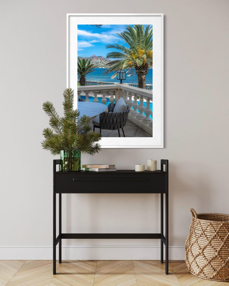 May include: A framed photograph of a scenic coastal view with palm trees, a pool, and a table and chair set. A black console table with a green vase and candles is in the foreground. A woven basket sits on the floor.