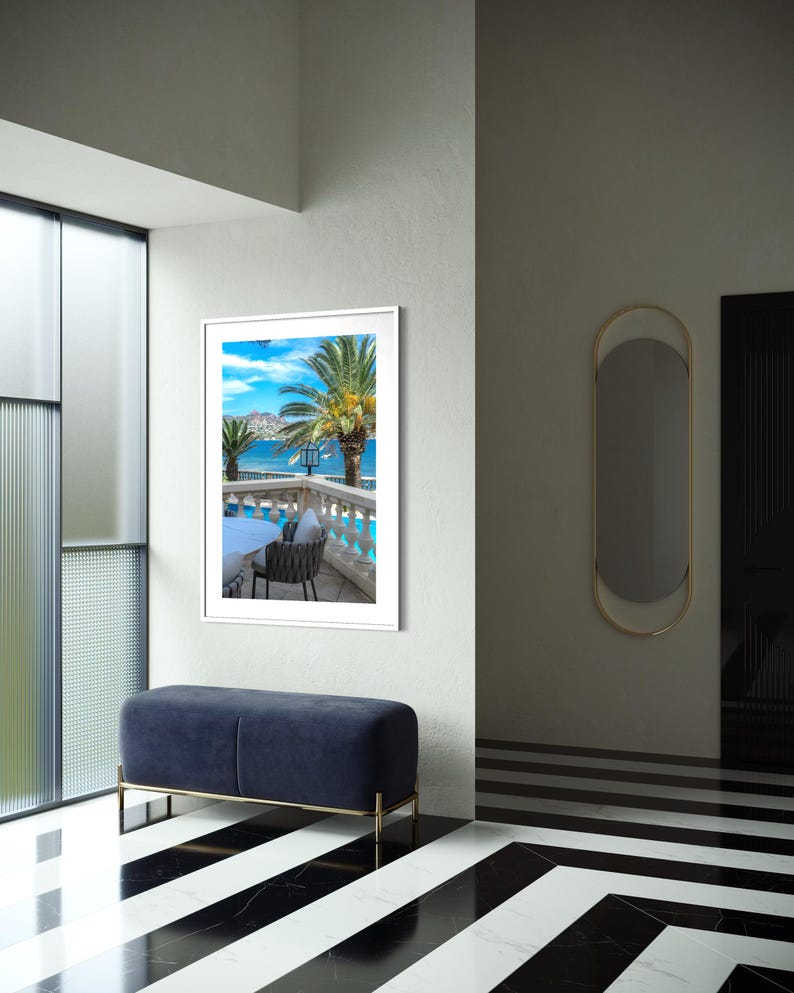 May include: An interior shot featuring a framed landscape print of a coastal scene with palm trees and a blue sky. A navy blue bench with gold legs sits below the artwork. The floor has a black and white striped pattern.