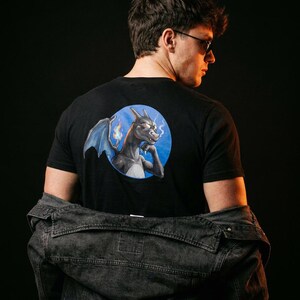 Könnte beinhalten: Ein schwarzes kurzärmeliges T-Shirt mit einer kreisförmigen Grafik auf der Rückseite. Das Motiv zeigt einen dunklen Drachen mit Sonnenbrille, blauen Flügeln und einer kleinen Flamme. Eine dunkelgraue Jeansjacke liegt über den Schultern.