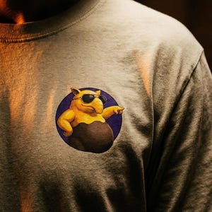 Puede incluir: Una camiseta de color beige claro con un gráfico circular en el pecho. El gráfico muestra un oso hormiguero amarillo con gafas de sol negras, señalando con una mano mientras apoya la otra en la cadera, sobre un fondo de color morado sólido.