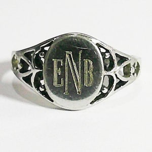 May include: Silver-toned ring with an oval face featuring the engraved initials "ENB" in a serif font. The ring has an openwork design with floral-like patterns on the band, creating a delicate and ornate appearance. The ring is set against a white background.