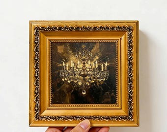May include: A square oil painting depicting a glowing crystal chandelier with multiple lit candles, set against a dark, textured background. The artwork is encased in an ornate, metallic gold frame with intricate embossed scrollwork patterns.