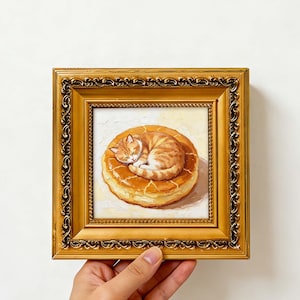 May include: A square oil painting in an ornate gold frame depicting an orange tabby cat curled up asleep on top of a round, golden-brown pastry. The text TIMBERDESIGNFINDS is printed in white at the bottom of the image.