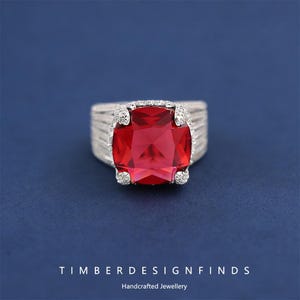 May include: A silver ring with a large, square, red gemstone. The ring has a textured band and small, clear accent stones around the red stone. The background is a solid blue. The text "TIMBERDESIGNFINDS Handcrafted Jewellery" is at the bottom.