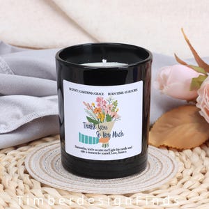 Puede incluir: Una vela de vidrio negro con una etiqueta blanca que presenta un ramo de flores y el texto "Thank You So Very Much". La etiqueta también incluye la fragancia "Gardenia Grace" y un tiempo de combustión de 45 horas. La vela está sobre un posavasos tejido.