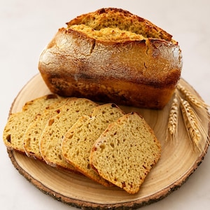 May include: A loaf of golden-brown bread, partially sliced, rests on a wooden serving board. Several slices are arranged in front of the loaf, showcasing the bread's texture. Wheat stalks are placed on the side, enhancing the rustic presentation.