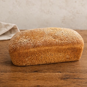 Könnte beinhalten: Ein rechteckiger Laib Vollkornbrot mit goldbrauner Kruste. Die Oberseite des Brotes ist leicht mit Mehl bestäubt und weist eine glatte, gebackene Textur auf. Der Laib ist horizontal ausgerichtet und zeigt seine gleichmäßige Form und Farbe.