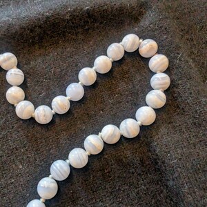 Può includere: Una collana composta da perline rotonde azzurre con venature bianche. Le perline sono infilate su un filo bianco, creando un gioiello delicato ed elegante. La collana è esposta su uno sfondo scuro e strutturato.