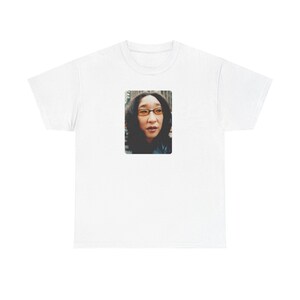 May include: A white short-sleeved t-shirt featuring a rectangular graphic print on the chest. The print displays a portrait of a person with dark hair wearing amber-tinted glasses, set against an urban background with blurred buildings.