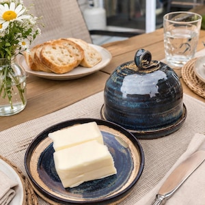Könnte beinhalten: Eine blaue Keramik-Butterdose mit passendem Deckel und ein Teller mit zwei Butterstücken. Die Dose hat eine dunkelblaue Glasur mit braunen Akzenten. Ein Teller mit Brot und ein Glas Wasser stehen auf dem Holztisch.
