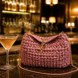 May include: A mauve crochet handbag featuring a textured knit pattern with embedded sequins, a gold-tone chain shoulder strap, and a gold-tone star-shaped charm hanging from the front closure, positioned next to a glass of light-colored liquid.