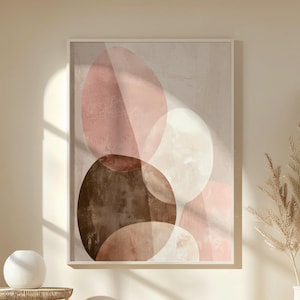 Puede incluir: Una impresión artística abstracta enmarcada con círculos superpuestos en tonos marrón, rosa y blanco. La obra de arte cuelga sobre un otomán blanco y una pequeña mesa auxiliar de madera. También se ve un jarrón con plantas secas.