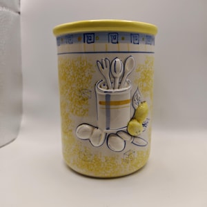 Può includere: Un portautensili in ceramica con un design maculato giallo e bianco. Il bordo superiore è giallo e il corpo presenta un bordo blu e bianco. Un design a rilievo include utensili, limoni e foglie. Questo articolo da cucina è ideale.