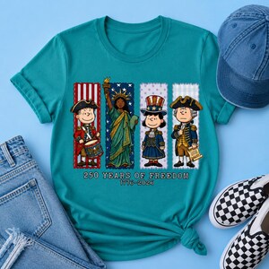 May include: A teal short-sleeve t-shirt featuring a graphic of four cartoon characters dressed in historical American attire, including the Statue of Liberty. The text reads 250 YEARS OF FREEDOM 1776-2026, set against vertical red, white, and blue stripes.