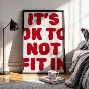 May include: A framed print with the text "IT'S OK TO NOT FIT IN" in red on a white background. The black-framed print is propped against a wall. A black lamp, a book, and a mug are in front of the print.