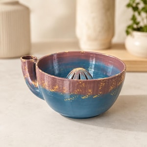 May include: A ceramic citrus juicer bowl featuring a deep blue base and a mauve upper rim with gold-toned speckling. The bowl includes a central reamer dome and a small pouring spout on the side, showcasing a hand-applied glaze finish.