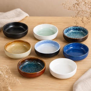 Op de afbeelding: Een verzameling van acht kleine, ronde keramische kommen in verschillende kleuren, waaronder zwart, wit, blauw en bruin. De kommen zijn gerangschikt op een licht houten oppervlak, waarbij hun verschillende texturen en afwerkingen worden getoond. Ideaal voor het serveren van dips, sauzen of kleine porties.
