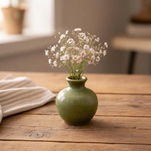 Op de afbeelding: Een kleine, ronde, groene keramische vaas met een boeket kleine, delicate, roze en witte bloemen. De vaas staat op een rustieke houten tafel. De vaas heeft een smalle hals en een bolvormig lichaam.