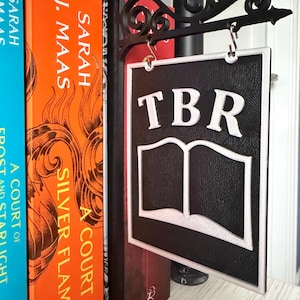 May include: A black and white TBR sign with an open book graphic hangs from a decorative black metal bracket. The sign is displayed near a stack of books with colorful covers. The sign's letters and book outline are white.