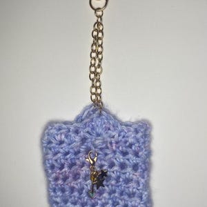 Puede incluir: Un estuche de ganchillo azul claro hecho a mano con una cadena y un cierre dorados. El estuche tiene un dije decorativo con un diseño de estrella y flecha. El trabajo de ganchillo tiene una apariencia texturizada, y el diseño general es caprichoso.