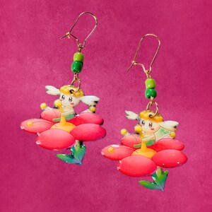 May include: A pair of dangle earrings featuring a cartoon character with white hair sitting on a large red flower with green leaves. Each earring has a gold-toned metal hook and a single bright green bead attached above the flower charm.