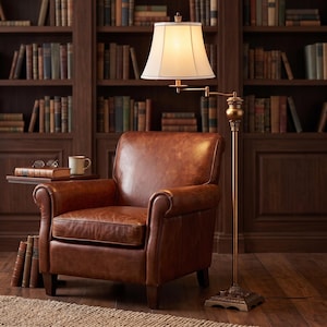 May include: A brown leather armchair with rolled arms, positioned next to a small side table holding a book, glasses, and a mug. A tall, antique-style brass floor lamp with a cream-colored fabric shade stands beside the chair on a wooden floor.