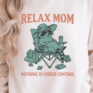 May include: Ivory-colored t-shirt with the text "RELAX MOM" in orange and "NOTHING IS UNDER CONTROL" in green. The shirt features a raccoon graphic wearing sunglasses and a hat, relaxing in a beach chair. The raccoon is holding a cup and is surrounded by beach accessories.