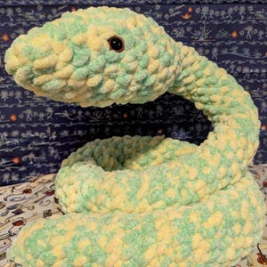 May include: A large, plush snake toy in shades of light green and yellow. The snake is coiled and has a round head with a dark eye. The soft, textured material gives it a cuddly appearance, perfect for a child's room or as a decorative item.