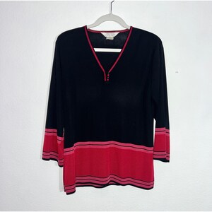 May include: A black long-sleeve pullover sweater featuring a V-neckline with red trim. The hem and sleeve cuffs are accented with wide horizontal bands of red and thin black stripes. The interior label displays the text Mirage.