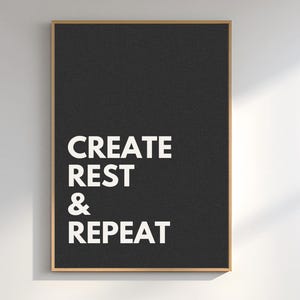 Puede incluir: Un póster negro enmarcado con las palabras "CREATE REST & REPEAT" en letras blancas en negrita. El póster tiene un diseño simple y minimalista y un marco marrón claro, adecuado para la decoración del hogar o la oficina.