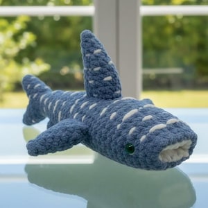 May include: A handmade blue and white crochet whale shark plush toy. The plush features white spots, a dorsal fin, and small side fins. The toy has a white mouth and green eyes. The plush is displayed on a reflective surface.