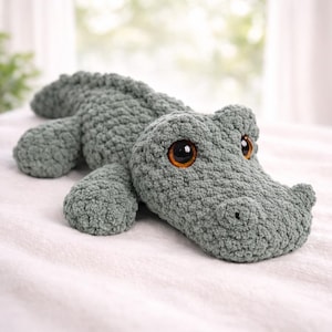 Puede incluir: Un peluche de cocodrilo hecho a mano, suave, de ganchillo, en un color verde apagado. El cocodrilo tiene ojos grandes, redondos, de color ámbar y una superficie texturizada y con bultos. El juguete está sobre una superficie blanca.