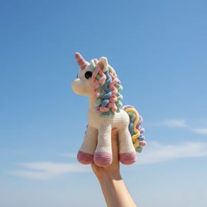 May include: A handmade crocheted unicorn toy. It has a cream-coloured body, pink hooves, and a pastel rainbow mane and tail. The unicorn features a pink horn and large black eyes. The toy is held against a bright blue sky.