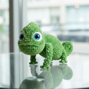 May include: A handmade green crochet chameleon with large blue eyes and white accents. The plush reptile has a rounded body, four small legs, and a curled tail. The chameleon is displayed on a reflective surface.