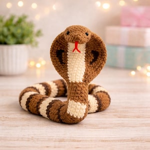 Könnte beinhalten: Ein gehäkeltes Kobra-Schlangenspielzeug in Braun-, Creme- und Weißtönen. Die Schlange hat eine erhobene Kapuze mit einem cremefarbenen Streifen in der Mitte und eine rote gespaltene Zunge. Der Körper ist mit Braun und Creme gestreift. Das Spielzeug liegt auf einer hellen Holzoberfläche.