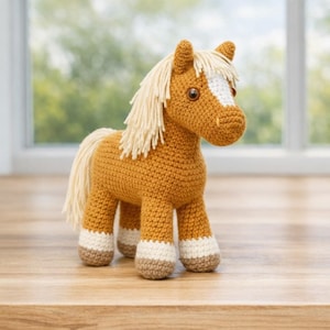 Puede incluir: Un juguete de caballo tejido a mano en tonos marrón, crema y beige. El caballo tiene una melena y cola de color crema, con una mancha blanca en la frente. Las pezuñas están detalladas con acentos crema y marrón. El juguete está sobre una superficie de madera.